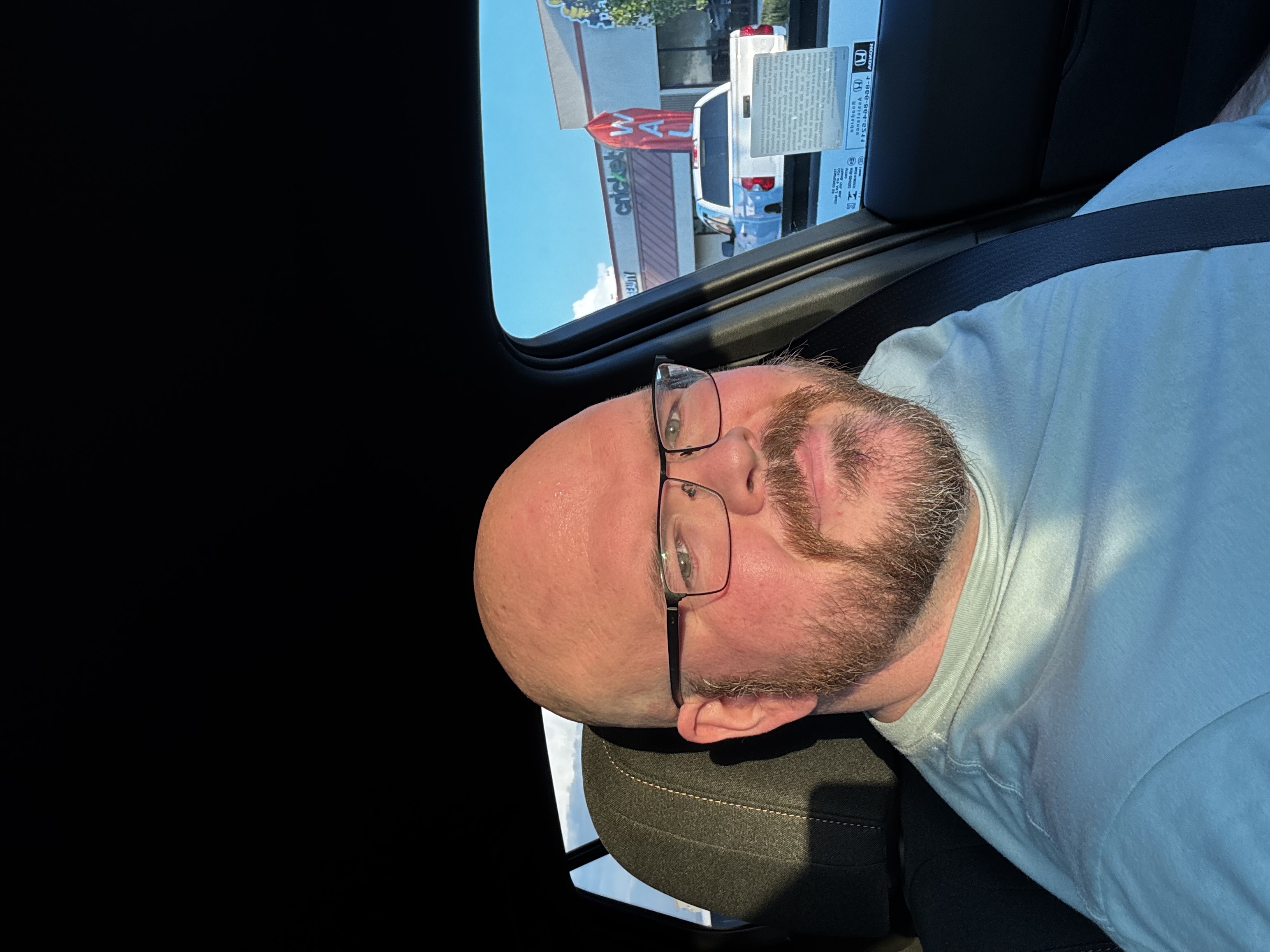 Sly Panorama selfie in the driver seat of a car wearing glasses and a seatbelt, daylight in a parking lot.