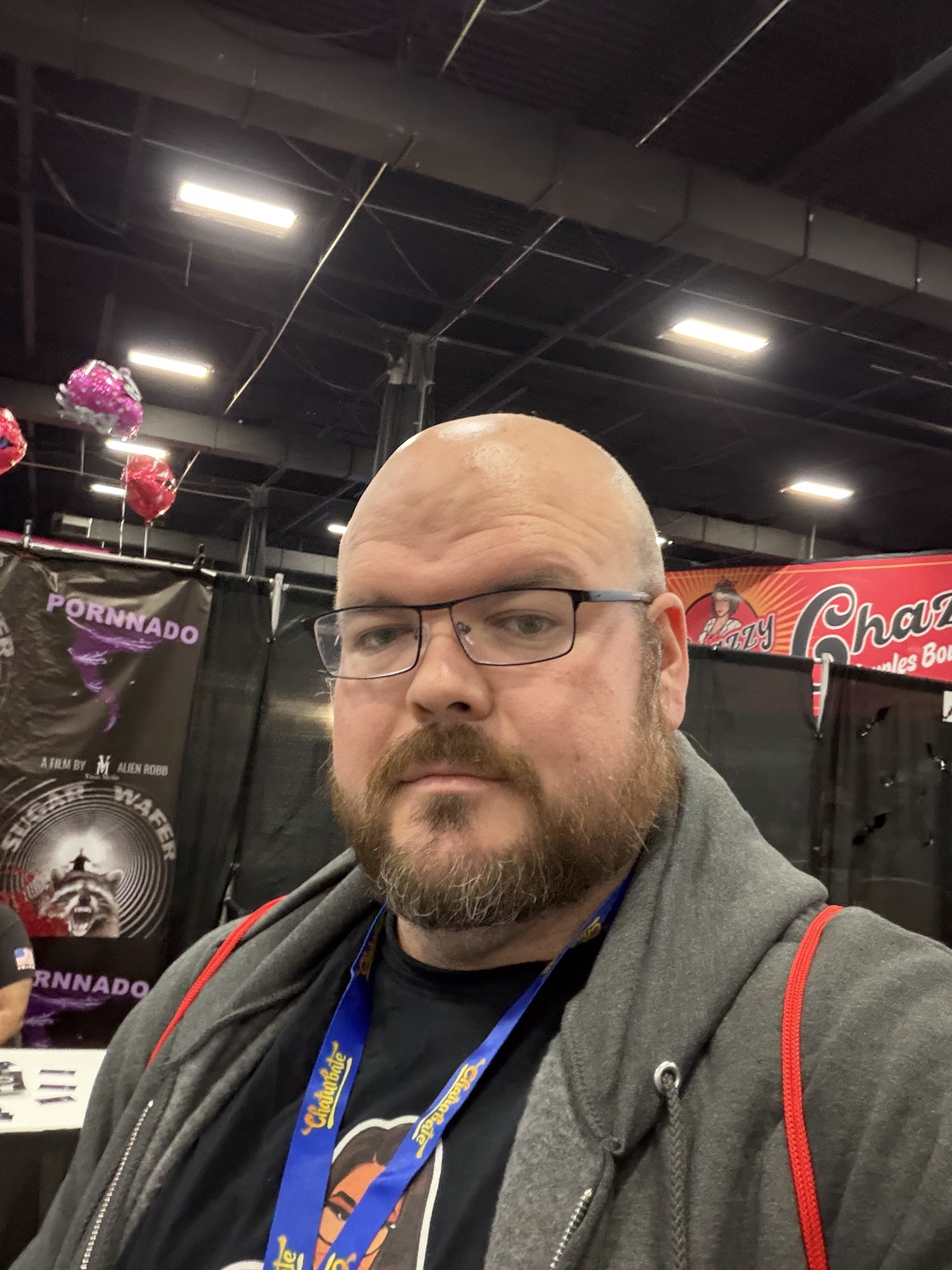 Sly Panorama at an adult industry convention wearing a Chaturbate lanyard and grey hoodie near expo banners.