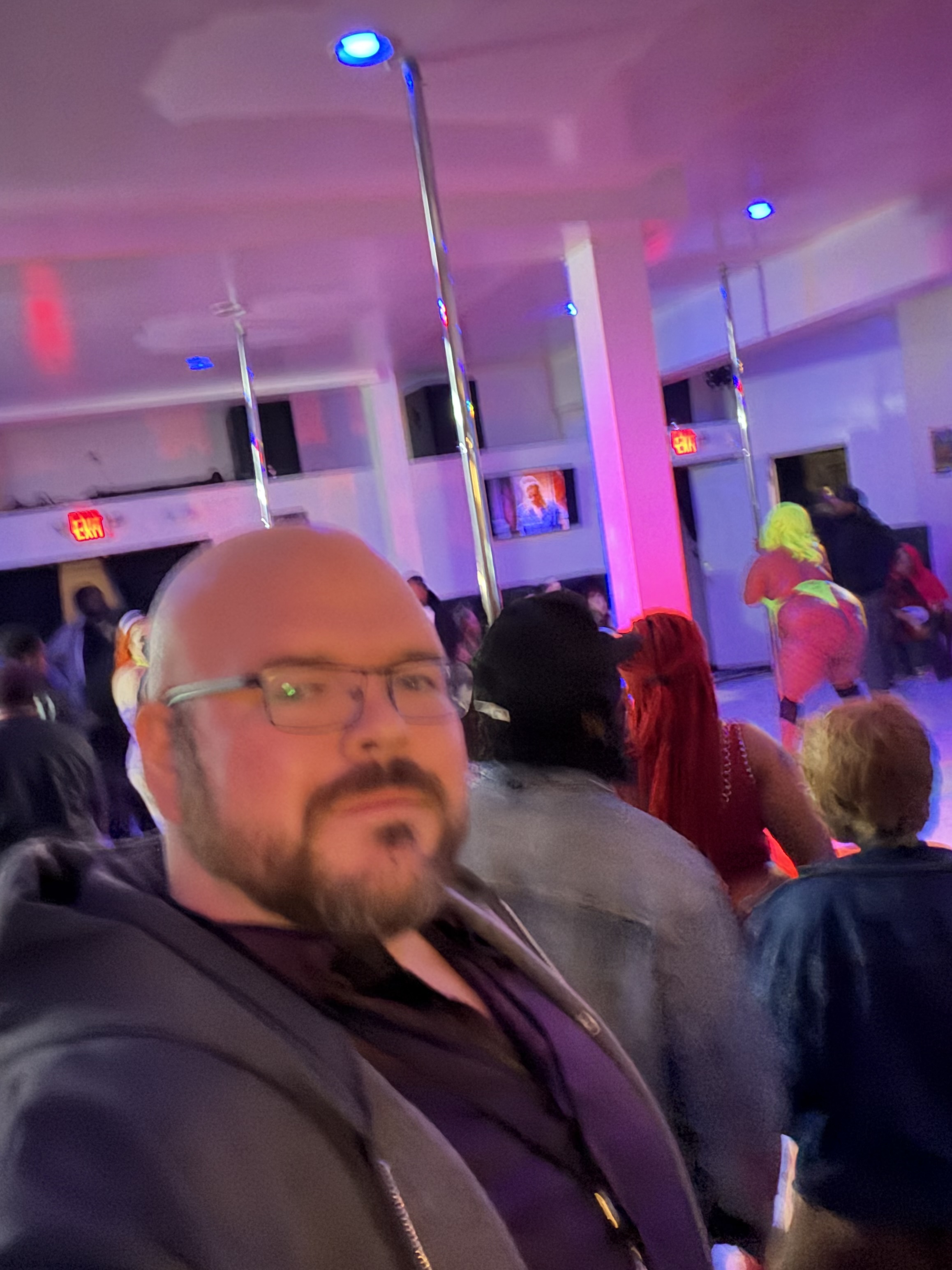 Sly Panorama selfie in a neon-lit nightclub with poles and colorful club lighting behind him.