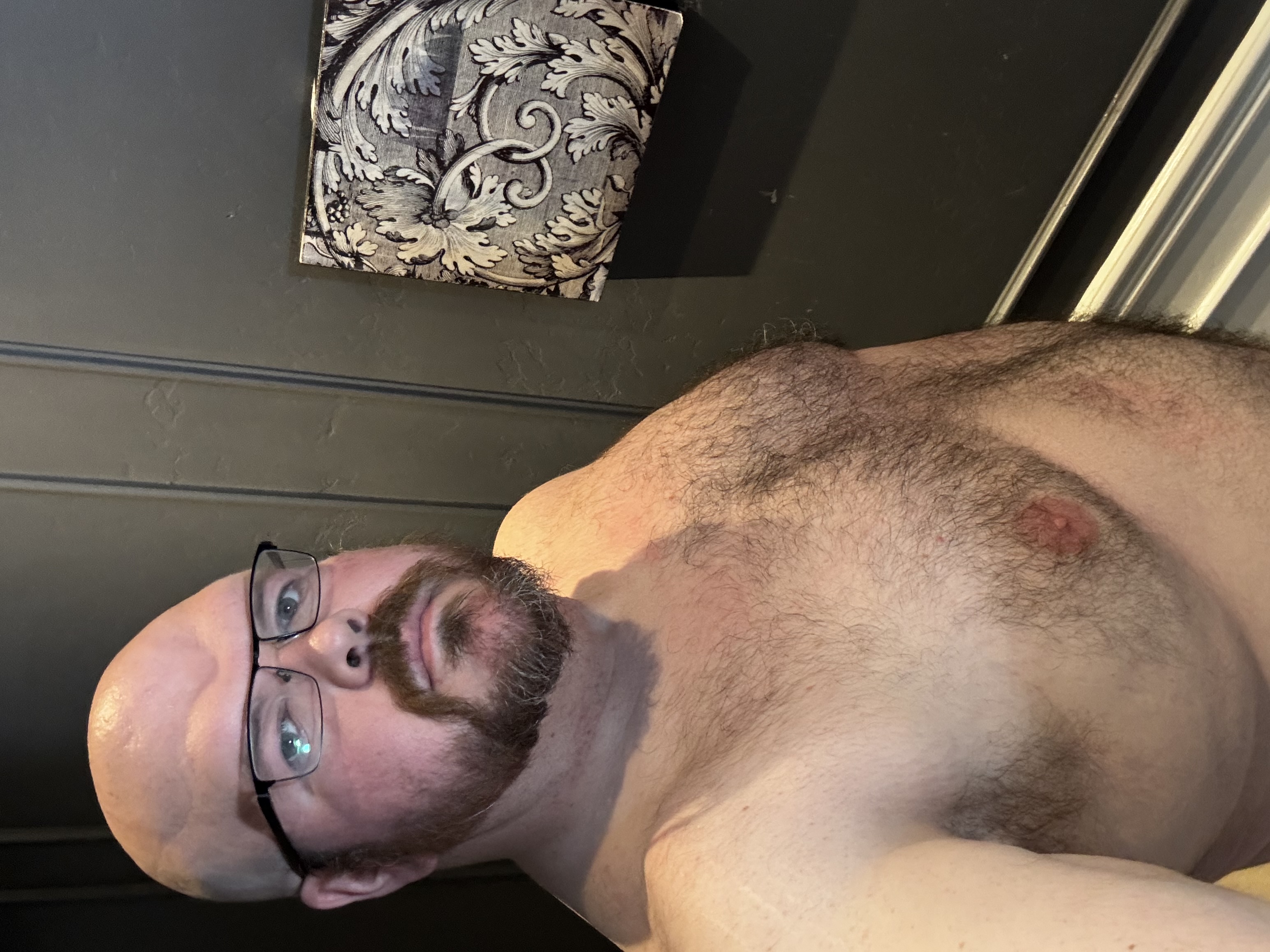 Sly Panorama shirtless selfie in front of a grey wall with framed damask-style art.
