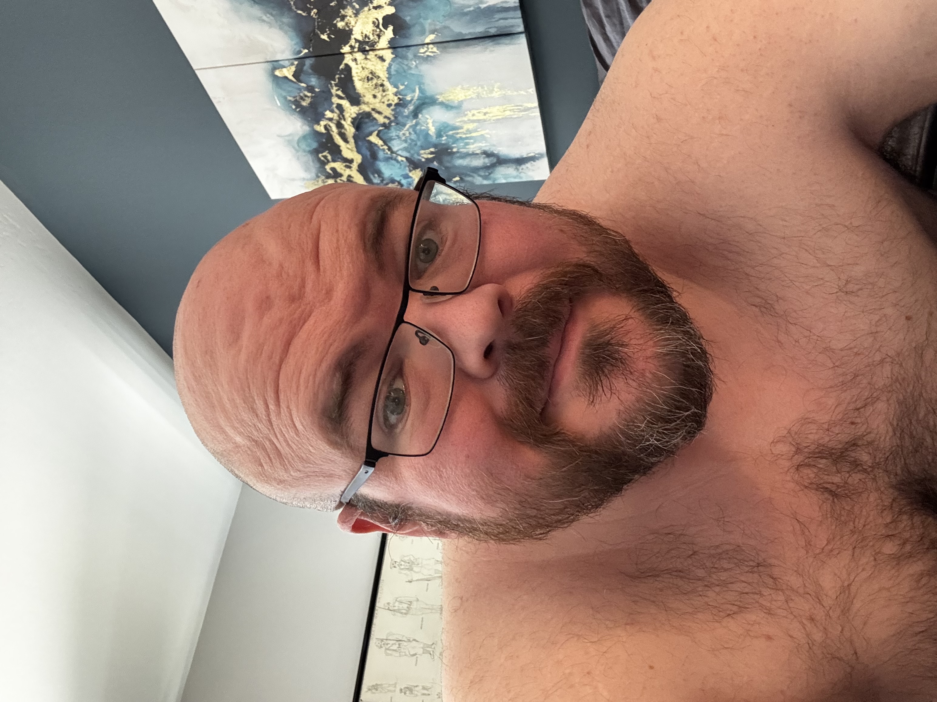 Sly Panorama shirtless selfie at home with blue wall and swirling blue, white, and gold artwork.
